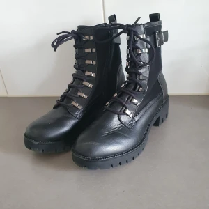 Svarta boots med snörning och spänne, deichman strl 38 - Tuffa svarta boots med grov sula, snörning framtill och coola metallhakar. Skorna har både skinn- och textildetaljer samt ett snyggt spänne vid ankeln. Perfekta för dig som vill ha en edgy stil med robust känsla.  Använda 2 gånger, lite nötta fram annars mycket fint skick