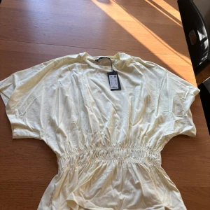 Ljusgul topp med resårmidja - Superfin ljusgul toppen från lager 157 med avslappnad passform och resår i midjan som ger en snygg siluett. Klänningen har korta ärmar och är gjord i mjuk bomull, perfekt för varma dagar. Enkel och stilren design som känns fräsch och modern.