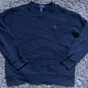Mörkblå sweatshirt från Polo Ralph Lauren - Klassisk mörkblå sweatshirt från Polo Ralph Lauren med röd broderad logga på bröstet. Tröjan har rund halsringning, långa ärmar och är gjord i mjuk bomull. Perfekt för en chill och stilren look.