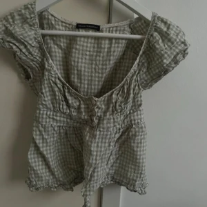 Grönvit rutig blus Brandy Melville - Supersöt rutig blus från Brandy Melville i grön och vitt. Modellen har korta puffärmar, v-ringning, knappar framtill och smock i midjan. Sömmen har spruckit i ”armhålan” på båda sidorna, säljer därför mycket billigt. 