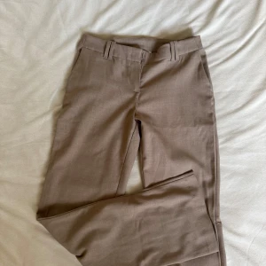 Beige kostymbyxor med bootcut - Snygga beige kostymbyxor med låg midja och bootcut ben. Byxorna har hällor för bälte, dold dragkedja och knappstängning i midjan. Perfekta för en clean och stilren outfit🦋. Materialet känns mjukt och har en lätt struktur.