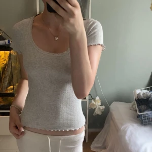 Ljusgrå ribbad topp från h&m - Supersöt ljusgrå ribbad topp från Divided med korta ärmar och söt spetskant vid halsringningen. Toppen har vågig kant nertill och vid ärmslut, samt en tight passform som sitter snyggt. Perfekt till jeans eller kjol för en chill look.