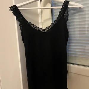 Svart spetslinne från Vero Moda - Svart linne från Vero Moda med breda axelband och v-ringning, dekorerat med snygg spets längs ringningen och axelbanden. Mjuk och stretchig känsla. Ny med lapp kvar.