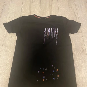 Svart AMIRI t-shirt med drip-print - Svart t-shirt från AMIRI med stort tryck av loggan framtill och baktill, där bokstäverna har färgglada drip-effekter i lila, blått och orange. T-shirten har korta ärmar och är gjord i mjuk bomull. Snygg detalj med färgstänk nedtill på framsidan. Tröjan är i storleken S. Pris kan diskuteras! 