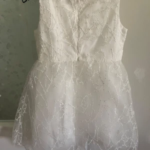 Vit kort klänning med rosett - Superfin vit kort klänning med glittriga broderade detaljer och en stor söt rosett i midjan. Klänningen är ärmlös och har ett skirt lager ovanpå som ger extra festlig känsla. Perfekt för speciella tillfällen när man vill känna sig extra fin.