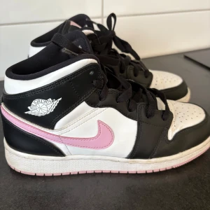 Nike Air Jordan 1 Mid GS Arctic Pink - Snygga Nike Air Jordan 1 sneakers i svart, vitt och rosa. Klassisk high-top modell med svart snörning, perforerad tå och rosa swoosh. Skorna är tillverkade i skinn och har en ikonisk Jordan-logga på sidan. Strl 39. Använda 10-20 ggr.