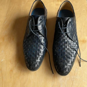 Svarta flätade skinnskor Giorgio 1958 - Snygga svarta skinnskor från Giorgio 1958, handgjorda i Italien. Skorna har ett unikt flätat mönster och klassisk snörning. Perfekta för dig som vill sticka ut med stilrena detaljer och exklusiv känsla. Skorna har rund tå och låg klack.