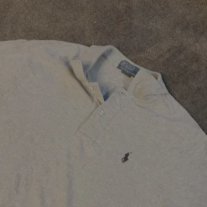 Pikétröja från Polo Ralph Lauren - Klassisk ljusgrå pikétröja från Polo Ralph Lauren med broderad logga på bröstet. Modellen är 173 cm och den sitter lite långt på han. Tröjan har korta ärmar, krage och knäppning framtill. Tillverkad i 100% bomull för en skön och luftig känsla. Perfekt till jeans eller shorts. Pris kan diskuteras och hesitera inte att fråga om någon beundran