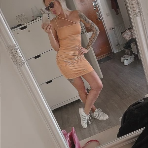 Persikofärgad tight kortklänning - Säljer en snygg persikofärgad kortklänning med asymmetrisk axel och smala axelband. Klänningen är figurnära och har rynkade sidor som ger en cool effekt. Perfekt för dig som gillar trendiga och stilrena plagg. Klänningen har en fläck i sidan av Klänningen som ej går bort.