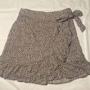 Blommig brun omlottkjol från BikBok - Brun kort omlottkjol från BikBok i blommigt mönster. Kjolen har volangkant nertill och knyts i sidan med en rosett. 