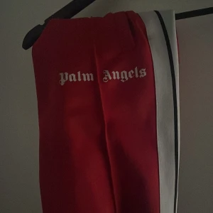 Palm Angels röda trackpants XS - Riktigt snygga röda trackpants från Palm Angels med vit och svart sidoremsa. Byxorna har elastisk midja och logga framtill. Tillverkade i 100% polyester för en sportig vibe. Perfekta för dig som vill sticka ut med streetwear.