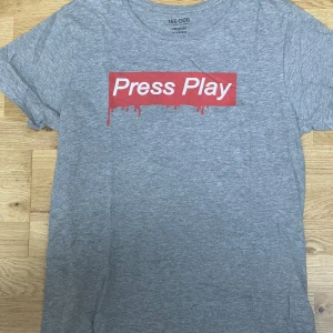 Grå t-shirt Press Play 157.DOG - Gråmelerad t-shirt från 157.DOG med röd tryckt ruta och texten 'Press Play' på bröstet. Klassisk rund hals och korta ärmar. Skön och mjuk bomullskänsla, perfekt till jeans eller shorts. Snyggt street-inspirerat tryck som sticker ut.