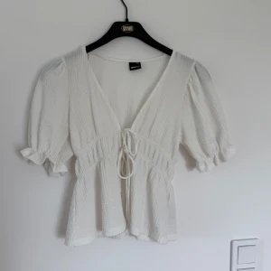 Vit blus med knyt och puffärm - Supersöt vit blus från Gina Tricot med v-ringning, knyt framtill och puffiga korta ärmar med volangkant. Blusen har en lätt strukturerad bomullskänsla och ger en romantisk vibe. Perfekt att styla med jeans eller kjol.