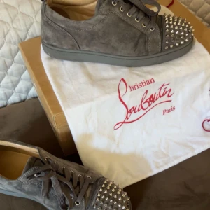 Grå mocka sneakers från Louboutin - Storlek 42, 2 stycken nitar saknas men det finns 2 extra som man smidigt kan sätta på. Mockan är i väldigt bra skick.