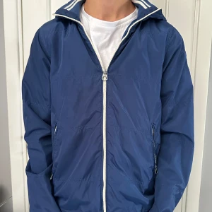 SCOTCH & SODA Jacka  - Snygg blå vindjacka med dragkedja och huva. Jackan har ribbade detaljer vid kragen och är perfekt för blåsiga dagar. Två praktiska fickor med dragkedja framtill.