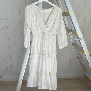Vit långklänning från Melmelli - Superfin vit långklänning från Melmelli med v-ringning och spetsdetaljer längs kanterna. Klänningen har trekvartsärm och är tillverkad i ett lätt bomullstyg, perfekt för varma dagar. Romantisk och flowy känsla med volanger och broderade detaljer.