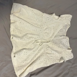 Vit ärmlös blus med broderade hål - Supersöt vit blus i bomull med broderade hål och romantiskt mönster. Toppen är ärmlös med volangkant och har en söt knytning framtill. Perfekt för varma dagar och ger en fräsch sommarkänsla.