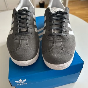 Adidas Gazelle grå/vita sneakers mocka - Snygga Adidas Gazelle sneakers i grå mocka med vita detaljer och klassiska tre ränder på sidorna. Skorna har vit sula, svart snörning och guldtexten 'GAZELLE' på sidan. Perfekta för dig som gillar retrostil och streetwear. De är bara använda 1 gång.