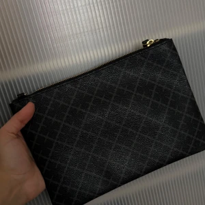 Svart kuvertväska från By Malene Birger - Stilren svart kuvertväska från By Malene Birger med diskret rutigt mönster och guldfärgad dragkedja. Väskan är i skinnliknande material och har ett ljusrosa innerfoder. Perfekt att bära i handen eller med den medföljande axelremmen. Använd endast ett fåtal gånger💕