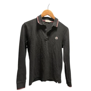 Mörkgrå Moncler pikétröja med logga - Snygg mörkgrå långärmad pikétröja från Moncler med klassisk krage och knäppning framtill. Kragen och ärmsluten har rödvita ränder och bröstet pryds av Monclers ikoniska logga. Perfekt för dig som gillar stilrena och sportiga plagg.