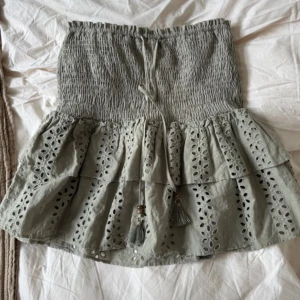 Grön volangkjol med broderade detaljer från Zara - Supersöt grå kortkjol med smock i midjan och knytband med tofsar. Kjolen har två lager volanger med broderade hålmönster som ger en romantisk vibe. Perfekt för varma dagar och lätt att matcha med en enkel topp. Inga defekter, inbyggda shorts. Stor i storlek passar S och m