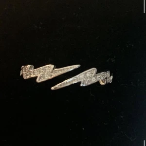 Blixtformade silverörhängen - Säljer ett par coola örhängen i form av blixtar, täckta med glittrande stenar. De är i silverfärg och har en edgy vibe som verkligen sticker ut. Perfekta för dig som vill ha något unikt och trendigt i öronen.