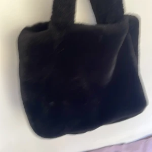 Svart fluffig handväska - Säljer en svart, fyrkantig handväska med mjuk och fluffig päls-look. Väskan har två korta handtag och en stilren design som sticker ut. Perfekt för dig som vill ha en mysig accessoar med trendig känsla. Är inte andvönd mycket 