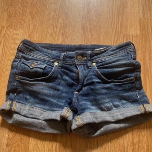 Blå jeansshorts från H&M, low waist - Snygga blå jeansshorts från H&M med låg midja och uppvikta benslut. Klassisk femficksmodell med coola detaljer som guldiga knappar och dekorativa sömmar. Perfekta för sommardagar och enkel att matcha med allt.
