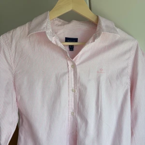 Rosa- och vitrandig skjorta från Gant - Klassisk skjorta från Gant i ljusrosa och vitt randigt mönster. Skjortan har lång ärm, knappar framtill och en broderad Gant-logga på bröstet. Tillverkad i mjuk bomull och har en normal passform med klassisk krage. I nyskick, nypris 1300kr. 🌸✨