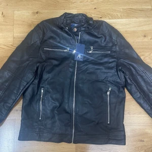 Svart skinnjacka från AC Made in Italy - Snygg svart skinnjacka från AC Made in Italy med flera dragkedjor framtill och på bröstet. Jackan har en klassisk bikerstil med ståkrage och detaljerade sömmar på axlar och ärmar. Perfekt för dig som vill ha en stilren och edgy look.