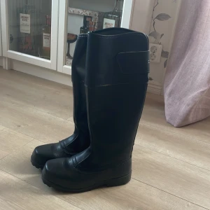 Svarta ridstövlar från Graninge - Svarta höga ridstövlar från Graninge med grov sula och kardborreknäppning upptill. Tillverkade i slitstarkt läder  med förstärkt tå och häl. Perfekta för dig som vill ha stabilitet och grepp i stallet.