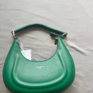 Grön axelväska från David Jones - Snygg grön axelväska från David Jones Paris med rundad form och guldfärgade metalldetaljer. Väskan har en bred axelrem och dragkedja upptill. Tillverkad i slätt syntetmaterial som ger en modern vibe. Perfekt för att lyfta din outfit med en pop av färg.