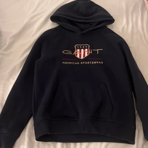 Mörkblå hoodie från GANT med tryck - Snygg mörkblå hoodie från GANT med broderat logotyp-tryck och sköld på bröstet. Klassisk känguruficka framtill och huva. Perfekt för dig som gillar sportig och stilren look. Mjuk insida i bomullsblandning. Säljer i storlek 158/164 (13-14år) men skulle säga att den passar xxs/xs också💞