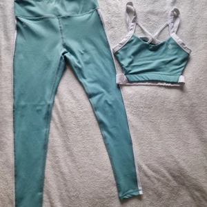 Workout Empire - Turkos och vit träningstopp och leggings - Snyggt set med turkos och vit träningstopp och matchande leggings. Toppen har korsade axelband och leggingsen har en hög midja med vita detaljer längs sidorna. Perfekt för gymmet eller yoga! Använd bara ett par gånger, det blev för liten för mig tyvärr.  