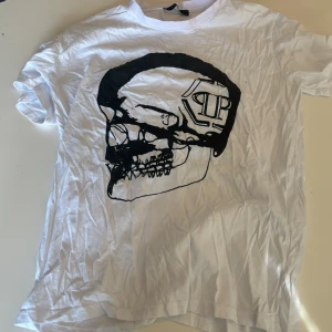 Filip Plain Vit t-shirt med dödskalletryck från Philipp Plein - Cool vit t-shirt från Philipp Plein med ett stort svart dödskalletryck på framsidan. Perfekt för dig som gillar en edgy stil. T-shirten är kortärmad och gjord i mjukt material för en bekväm passform.