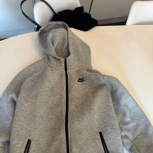 Grå hoodie från Nike - Säljer en grå hoodie från Nike med dragkedja och huva. Jackan har en enkel och stilren design med Nike-logga på bröstet. Perfekt för en avslappnad stil.