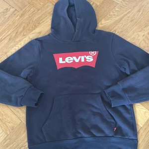 Mörkblå hoodie från Levi's - Säljer en mörkblå hoodie från Levi's med klassisk röd logga framtill. Tröjan har huva och en stor magficka. Perfekt för en avslappnad stil. Storlek XS/S