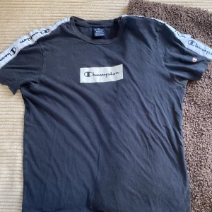 Svart t-shirt från Champion med logga - Svart t-shirt från Champion med vit logga på bröstet och loggoband längs axlarna. Klassisk passform och korta ärmar. Perfekt för en sportig och avslappnad stil.
