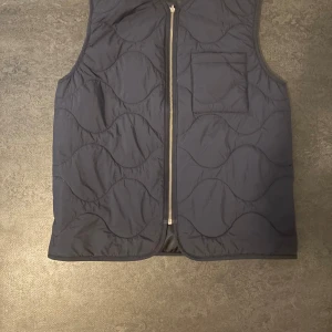 Mörkblå quiltad väst från Arket - Snygg mörkblå quiltad väst från Arket med dragkedja framtill och en bröstficka. Västen har ett vågigt mönster i quiltningen och är perfekt för lager-på-lager-stil.