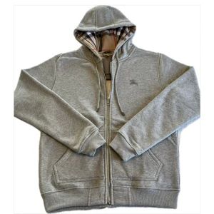 Grå hoodie från Burberry - Säljer en stilren grå hoodie från Burberry med klassiskt rutigt foder i huvan. Tröjan har dragkedja framtill och ribbade muddar vid ärmslut och nederkant. Perfekt för en avslappnad och trendig look.