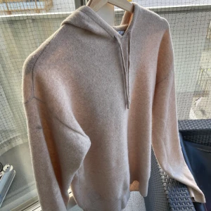 Beige Kaschmir hoodie från Carmés - Skön beige hoodie från Carmés i Kaschmir material. Tröjan har huva med dragsko och långa ärmar. Ribbstickade muddar vid ärmslut och nederkant ger en snygg passform.