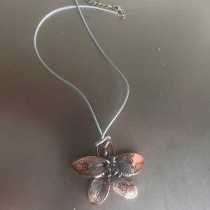 Halsband med blomma i kopparfärg - Unikt halsband med ett stort blomformat hänge i kopparfärg och svarta detaljer. Hängs på en tunn, ljus rem med justerbar kedja. Perfekt för dig som vill sticka ut med ett annorlunda smycke.