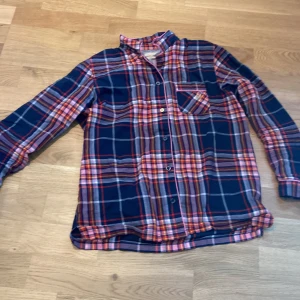 Rutig pyjamas skjorta till barn  - Snygg rutig skjorta från Holly & Whyte by Lindex i blått, rött och rosa. Skjortan har långa ärmar och en bröstficka med broderad detalj. Perfekt för en avslappnad stil.