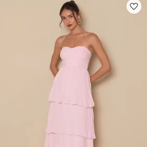 Rosa långklänning med volanger - Säljer en helt fantastisk ljusrosa långklänning med axelbandslös design och flera lager volanger. Klänningen har en figurnära passform upptill och faller vackert i långa, plisserade volanger. Perfekt för bal eller festliga tillfällen. Den är helt oanvänd med lappen kvar och säljes enbart på grund av att jag köpte 2 stycken! Dragkedjan är något svår att dra upp så därav jag dragit av några hundralappar från originalpriset (1400kr) om köparen skulle behöva byta ut dragkedjan. 