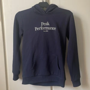 Mörkblå hoodie från Peak Performance - Mörkblå hoodie från Peak Performance med vit logotyp på bröstet. Klassisk huva och magficka framtill. Perfekt för en avslappnad stil och enkel att matcha med andra plagg. Storlek 160 JR original HOOD 