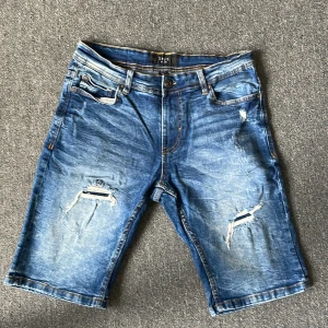 Blå slitna jeansshorts från SMOG - Säljer ett par blå jeansshorts från SMOG med slitna detaljer och klassisk femficksdesign. Shortsen har normal passform och är tillverkade i denim med snygga slitningar framtill. Perfekta för en avslappnad stil under varmare dagar.
