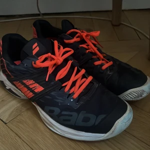 Svarta och orangea tennisskor från Babolat - Säljer ett par svarta tennisskor från Babolat med orangea detaljer och snören. Skorna har en robust sula och är designade för bra grepp på tennisbanan. Snygg logga på sidan och hälen. Perfekta för dig som vill ta spelet till nästa nivå.