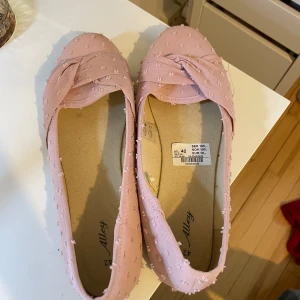 Ljusrosa ballerinaskor med rosett - Säljer ett par ljusrosa ballerinaskor från Ally med dekorativ rosett framtill och små prickiga detaljer i tyget. Skorna har rund tå och är tillverkade i syntetmaterial. Perfekta för dig som gillar en söt och feminin stil.