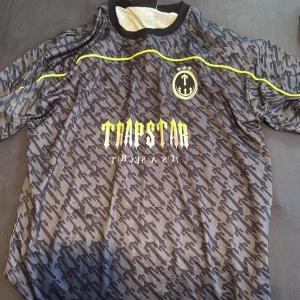 Trapstar svart fotbollströja med mönster och gula detaljer - Säljer en svart och grå fotbollströja från Trapstar med unikt mönster, gula detaljer och tryck. Tröjan har korta ärmar, rund hals och stort Trapstar-tryck både fram och bak samt siffran 22 på ryggen. Perfekt för dig som gillar streetwear och sportig stil. Passar perfekt inför sommaren! 