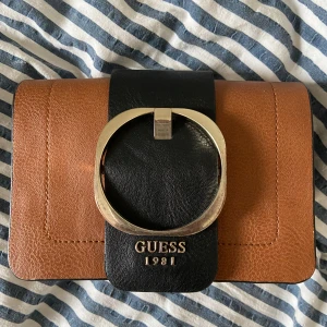  Väska från Guess - Säljer en snygg axelväska från Guess i brunt och svart skinn med stor guldfärgad ringdetalj framtill. Väskan har bred axelrem i beige och svart samt logga och årtal i guld. Har någon få defekt så skriv om du vill se det!